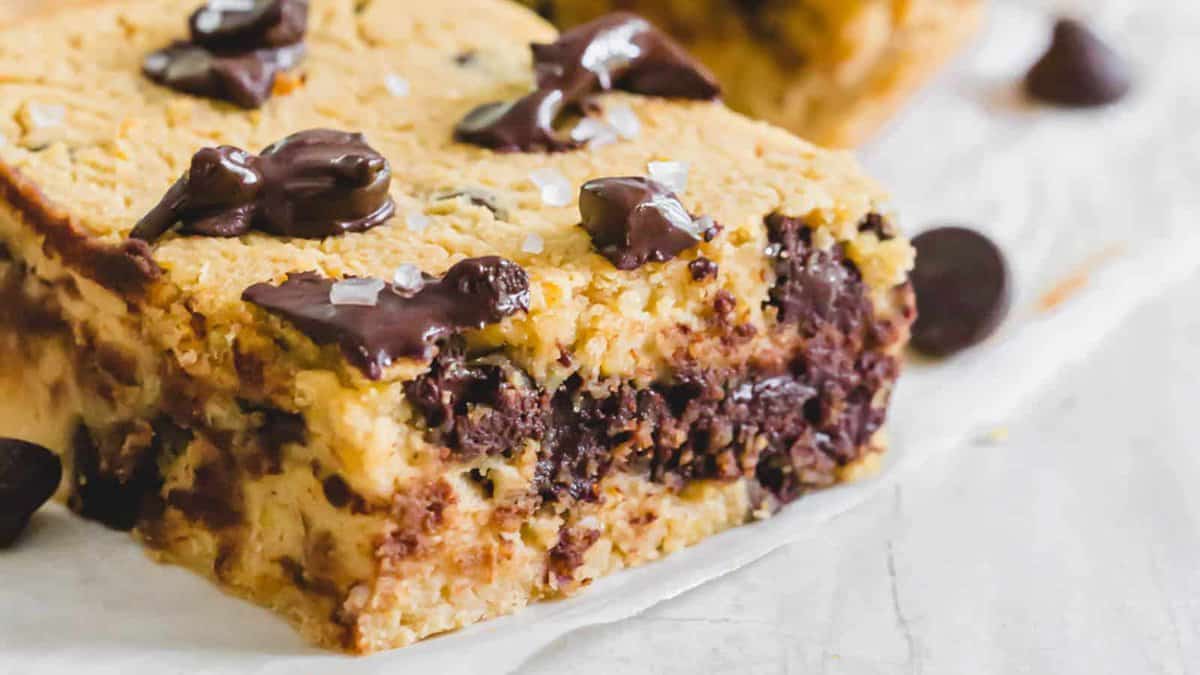 A close-up of a thick chocolate chip cookie bar with melted chocolate chunks and a sprinkle of sea salt on top, resting on parchment paper.