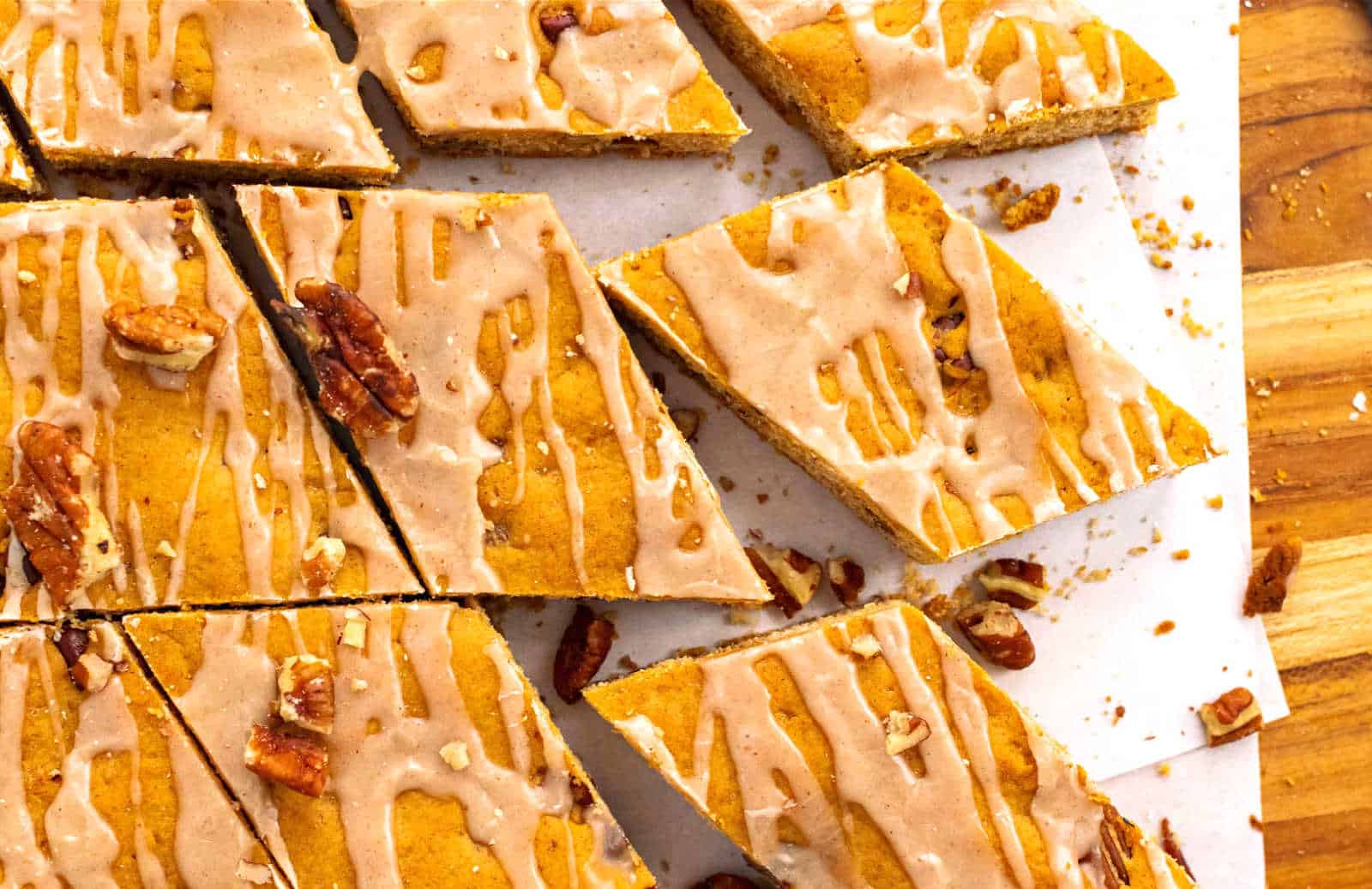 Pumpkin bars cut into triangles, topped with icing drizzle and pecans, arranged on parchment paper on a wooden surface.