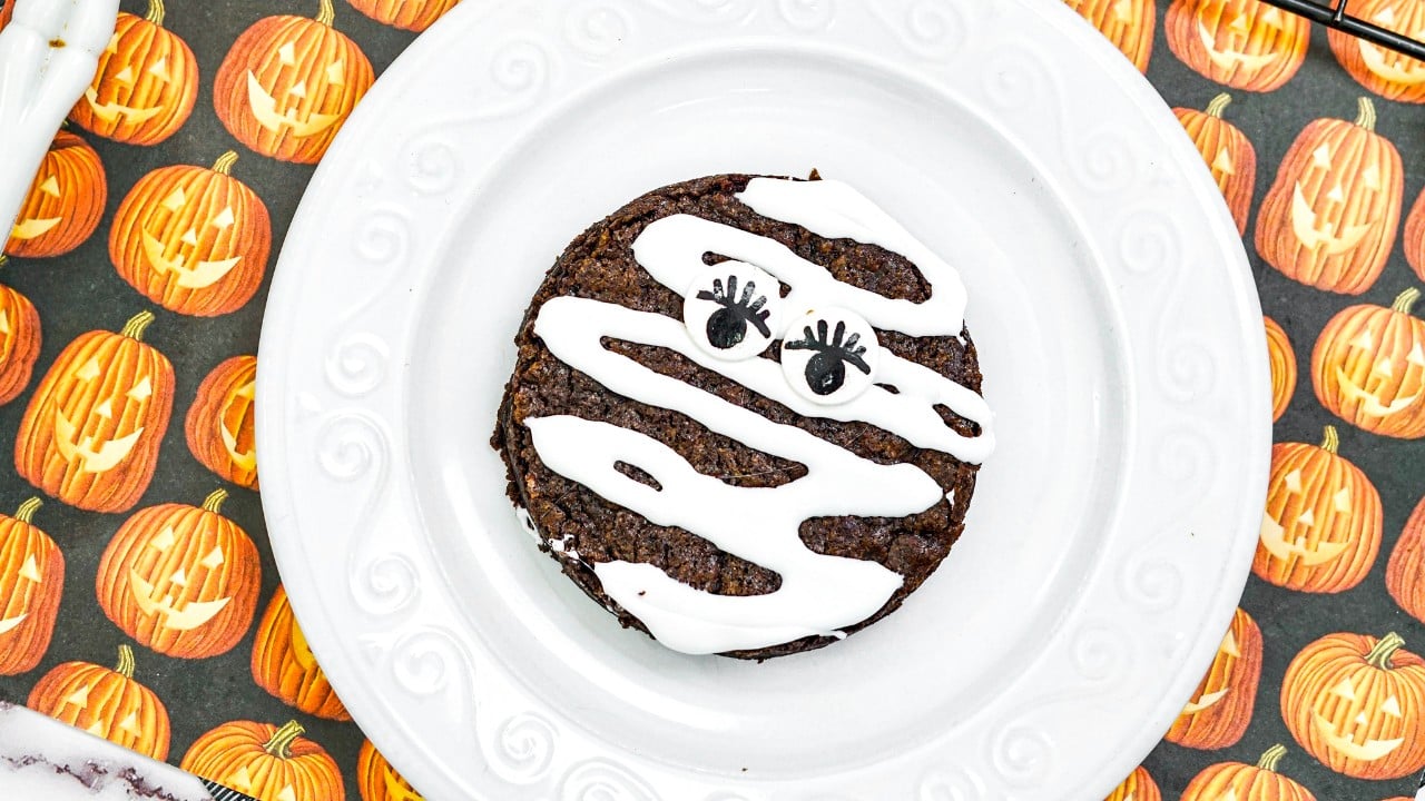 A chocolate cookie decorated with white icing and candy eyes to resemble a mummy, on a white plate with a Halloween pumpkin-themed background.