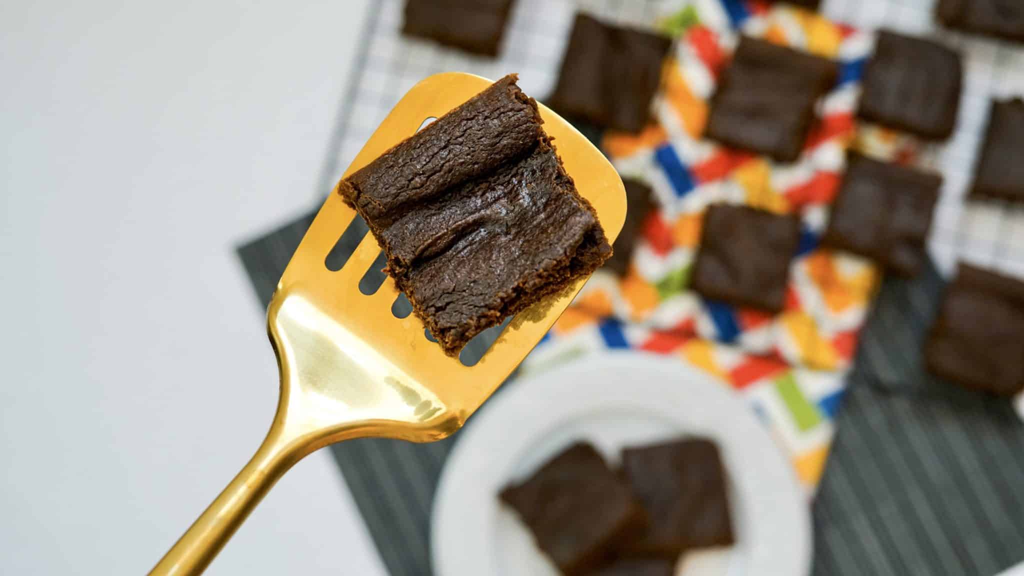A golden spatula holds a square piece of chocolate brownie above a plate and a colorful napkin with more brownies on a table.