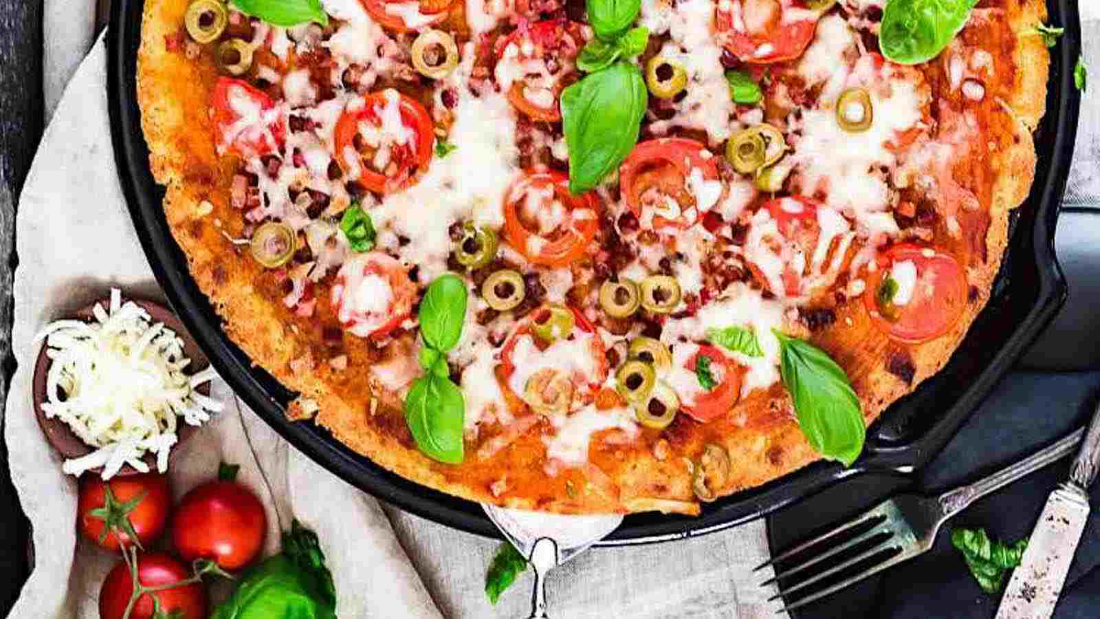 A baked pizza topped with melted cheese, sliced tomatoes, green olives, and fresh basil leaves, with a bowl of shredded cheese and tomatoes beside it.