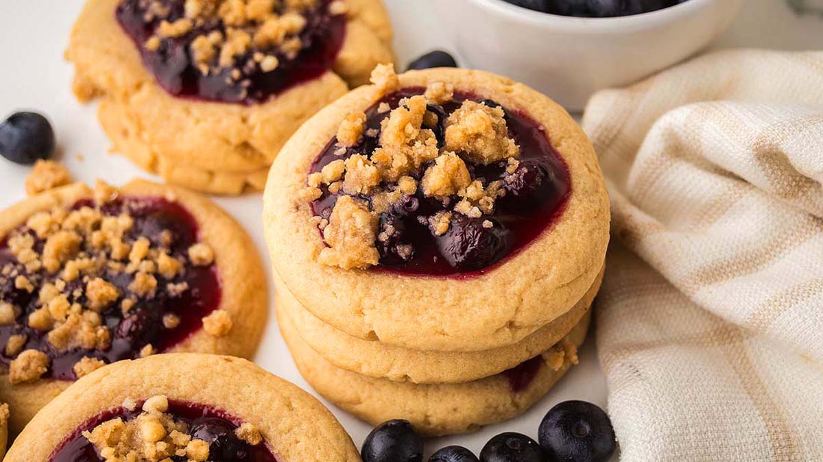 A stack of round cookies with a blueberry jam center and crumb topping, surrounded by fresh blueberries and a white napkin.