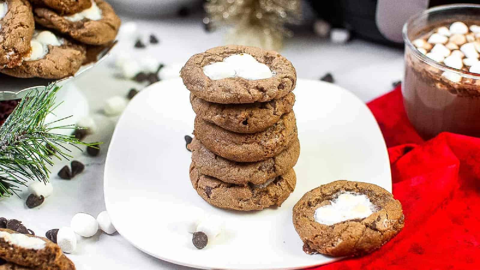 A stack of chocolate cookies with marshmallow centers is displayed on a white plate, surrounded by mini marshmallows and chocolate chips.