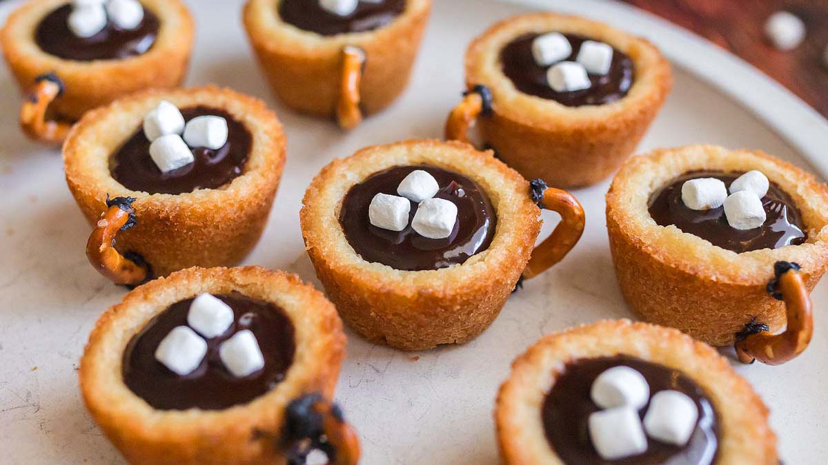 Mini cup-shaped cookies filled with chocolate and topped with small marshmallows, each with a small pretzel handle, arranged on a white plate.