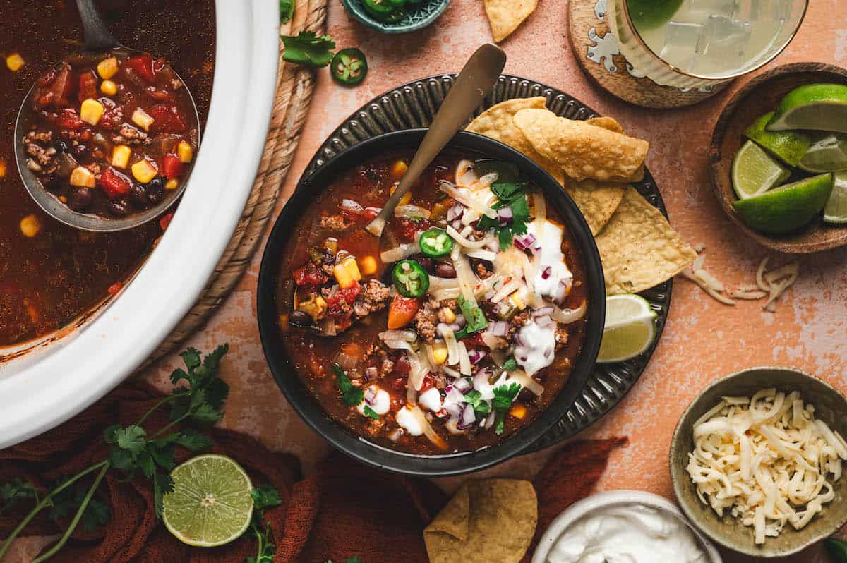 A bowl of chili topped with sour cream, shredded cheese, and jalapeรฑo slices, served with tortilla chips; a ladle of chili, lime wedges, and toppings are nearby.
