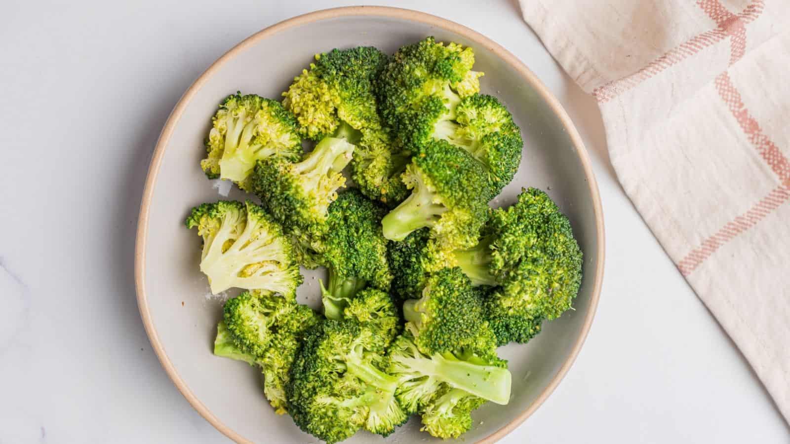 A bowl of steamed broccoli florets sits on a white surface next to a folded beige and pink plaid napkin.