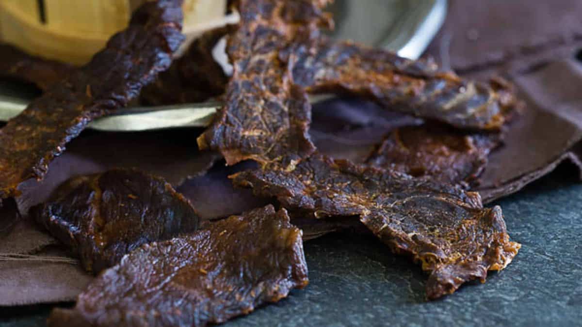 Several pieces of dark, dried beef jerky are arranged on a dark surface and brown cloth.