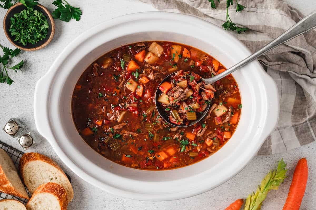 A white slow cooker filled with vegetable beef soup, with a ladle lifting a serving; bread slices, parsley, carrot, celery, and a towel surround it on a white surface.