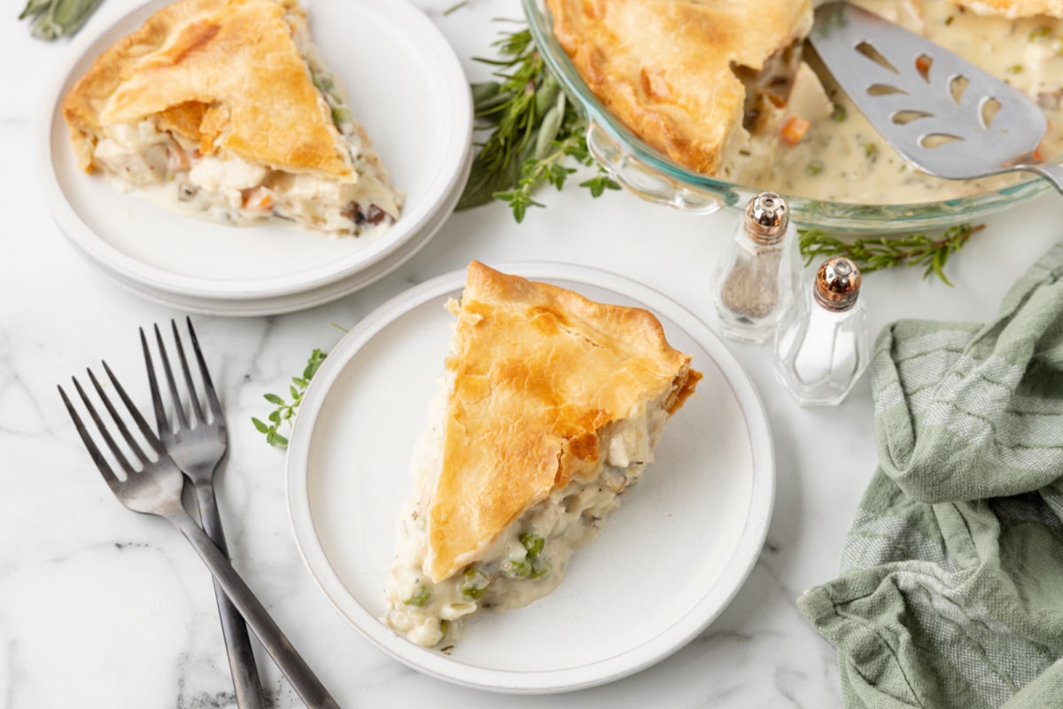 A slice of chicken pot pie with a golden crust sits on a white plate, with another serving, forks, herbs, and salt and pepper shakers nearby.
