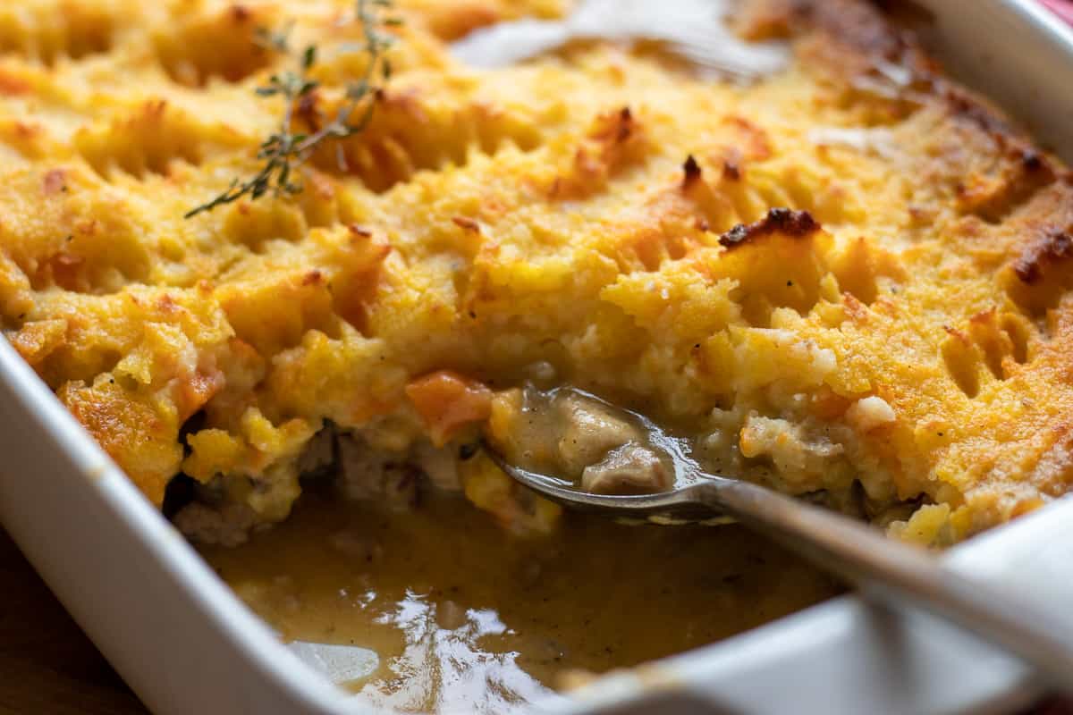 A close-up of a baked casserole with a golden mashed potato topping, a spoon scooping out a portion revealing a creamy filling beneath.