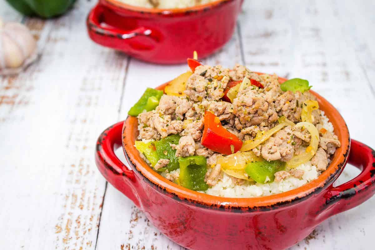 A red ceramic bowl filled with rice, ground meat, green and red bell peppers, and onions on a rustic white wooden surface.
