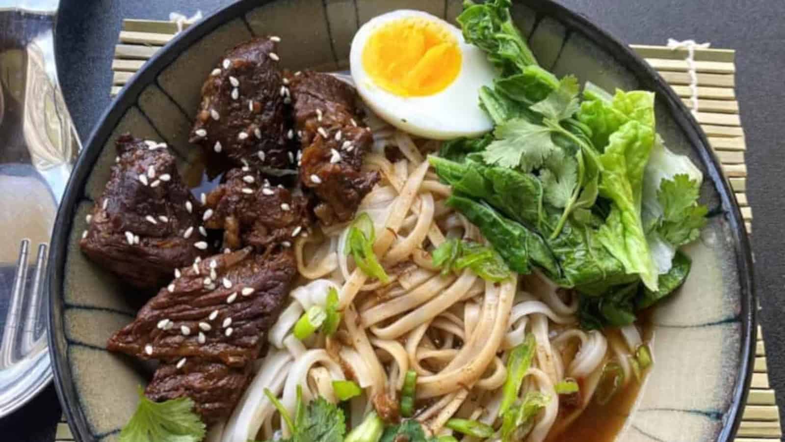 A bowl of noodle soup with beef pieces topped with sesame seeds, leafy greens, fresh herbs, and half a boiled egg.