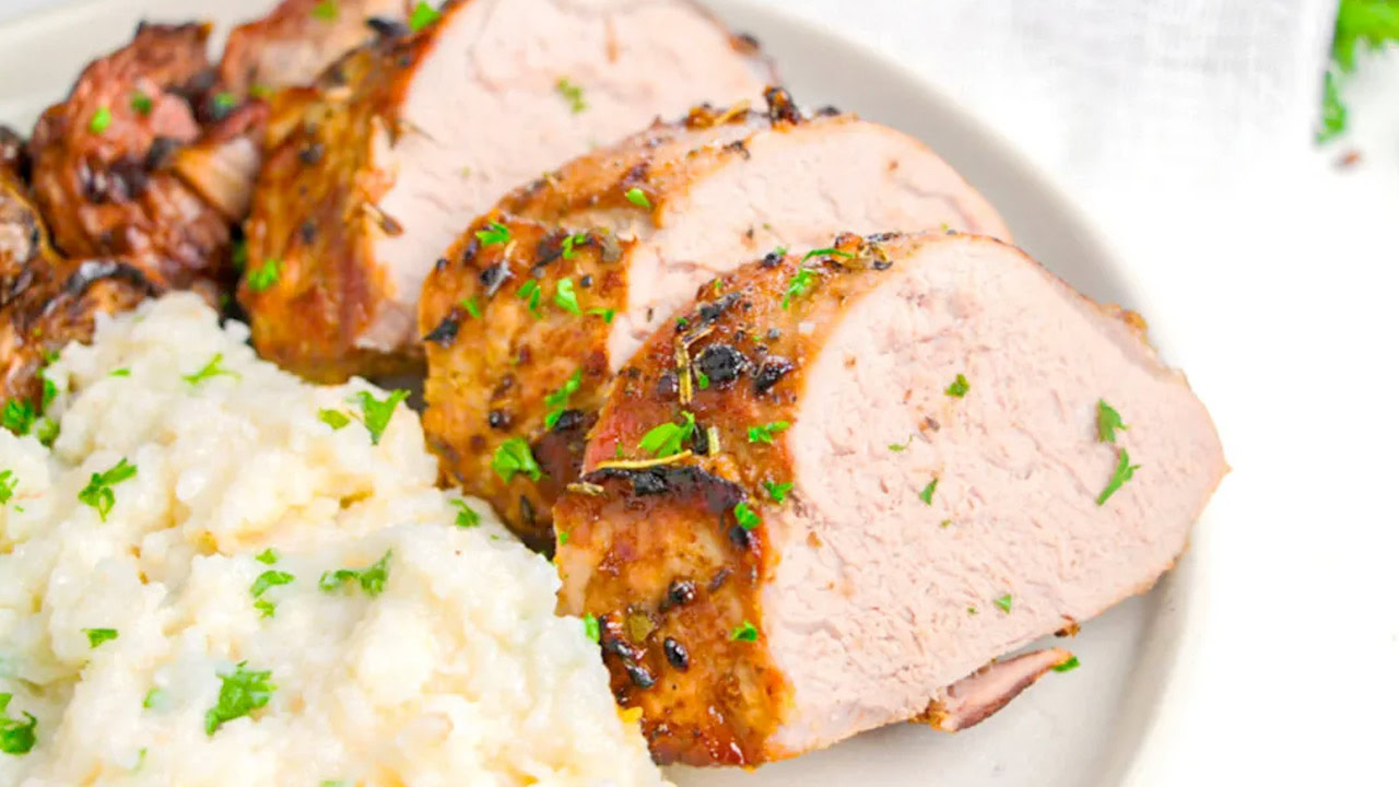 Sliced roasted pork tenderloin garnished with herbs, served next to a portion of mashed potatoes on a white plate.
