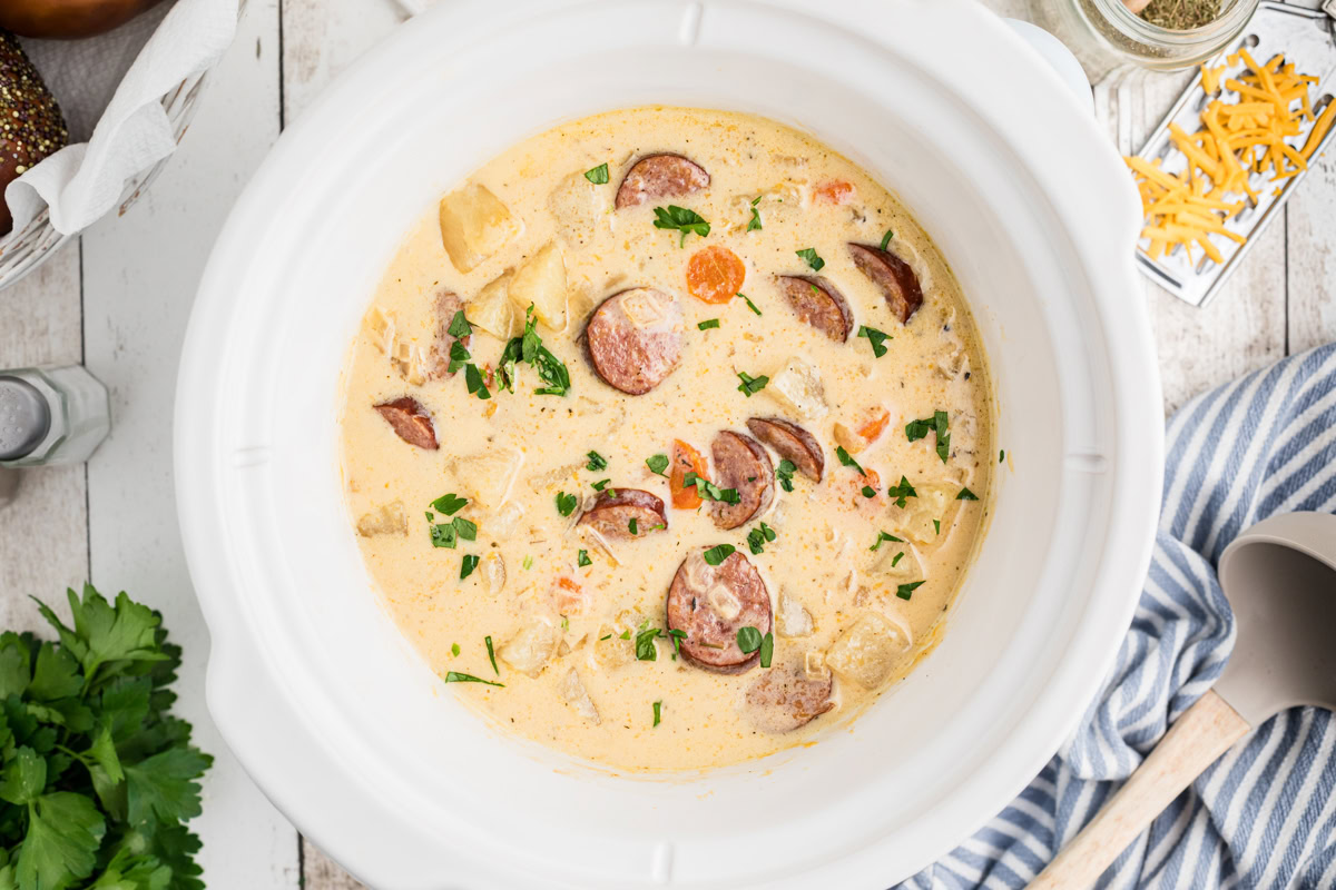A creamy sausage and potato soup with vegetables served in a white slow cooker, garnished with chopped parsley.