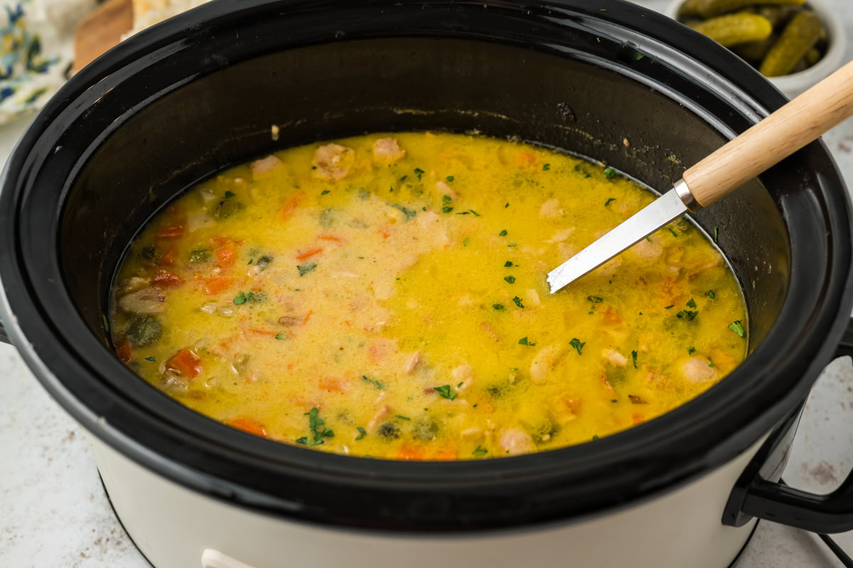 A crockpot filled with creamy, chunky soup containing visible pieces of vegetables and herbs, with a ladle resting inside.