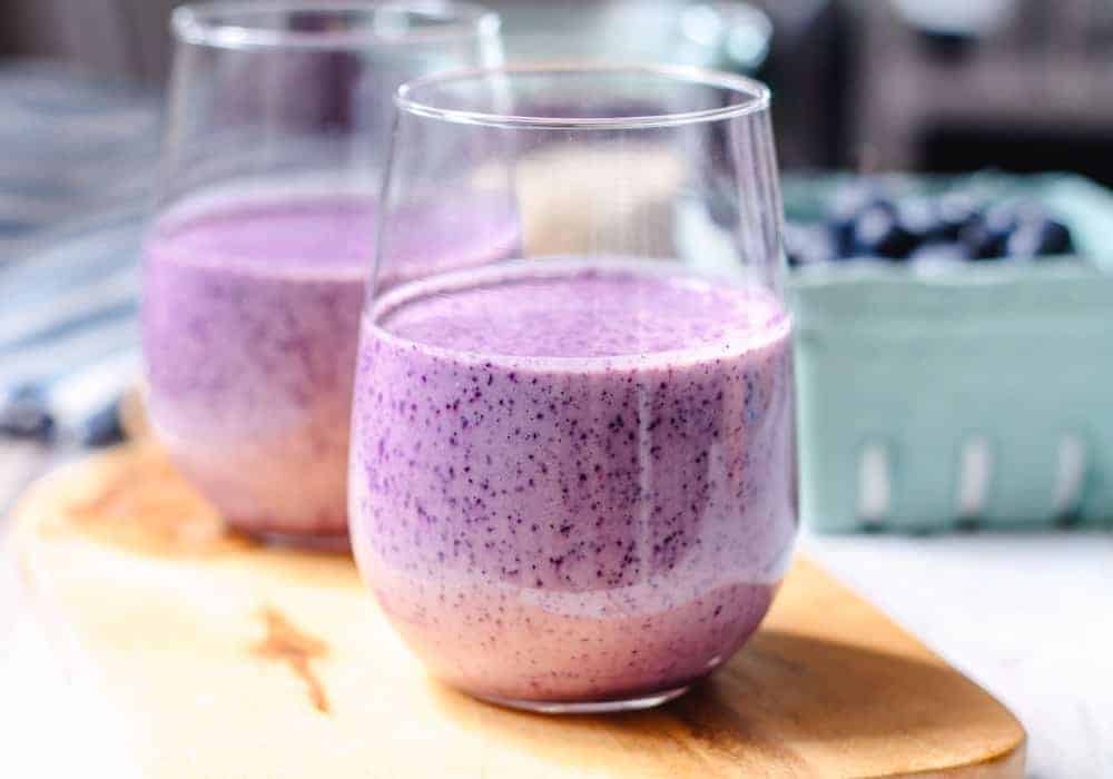 Two glasses filled with a purple blueberry smoothie sit on a wooden board, with a container of blueberries in the background.