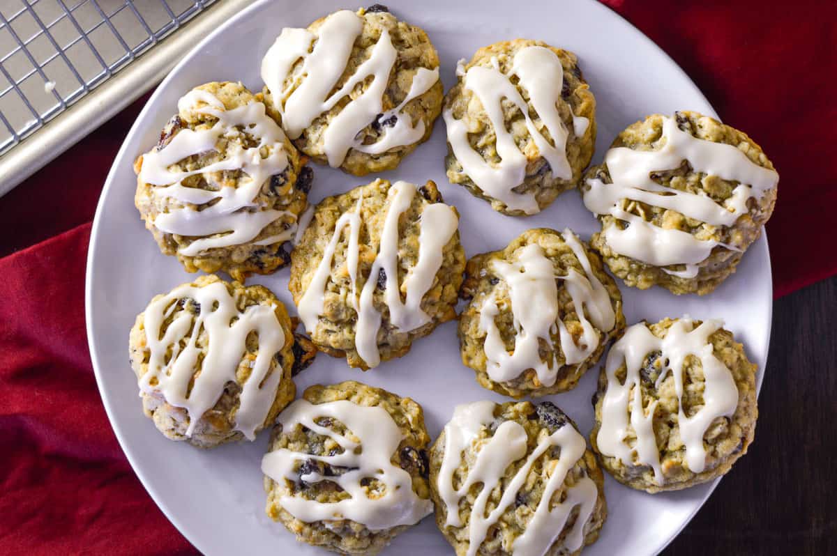 A white plate with nine oatmeal cookies topped with white icing drizzle, placed on a dark surface next to a red cloth and a cooling rack.