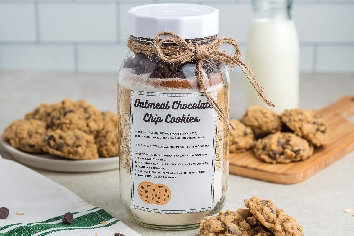 A jar labeled โOatmeal Chocolate Chip Cookiesโ filled with cookie ingredients sits on a counter with baked cookies, a plate, and a bottle of milk in the background.