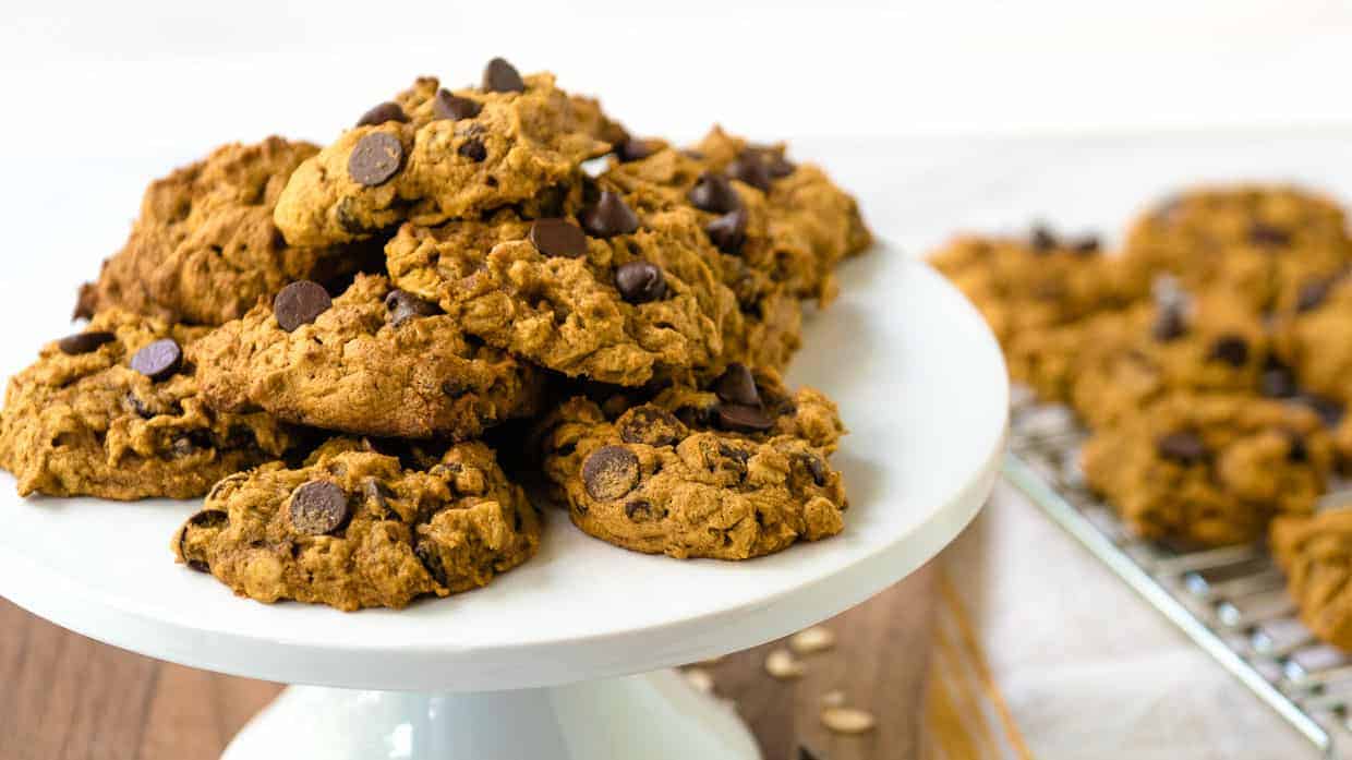 A pile of chocolate chip cookies is displayed on a white cake stand, with more cookies cooling on a wire rack in the background.