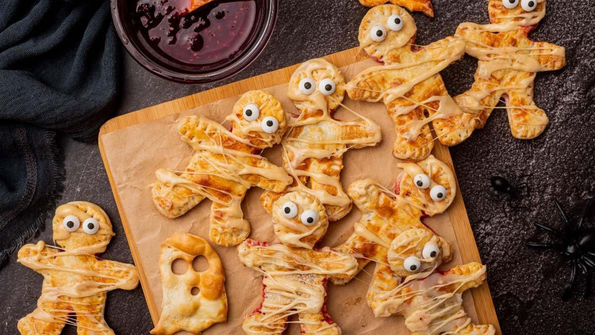 Baked pastries shaped like mummies with candy eyes and cheese wrappings on a wooden board, with a bowl of red dipping sauce nearby.