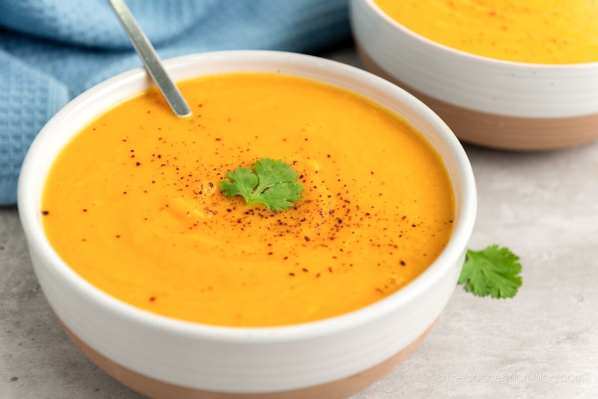 A bowl of creamy orange soup garnished with a cilantro leaf and sprinkled with pepper, with a spoon resting inside. Another bowl of soup and a blue towel are in the background.