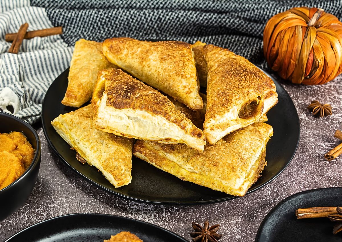 A black plate holds several golden-brown, triangular puff pastry turnovers. Decorative items and a bowl of filling are visible nearby.