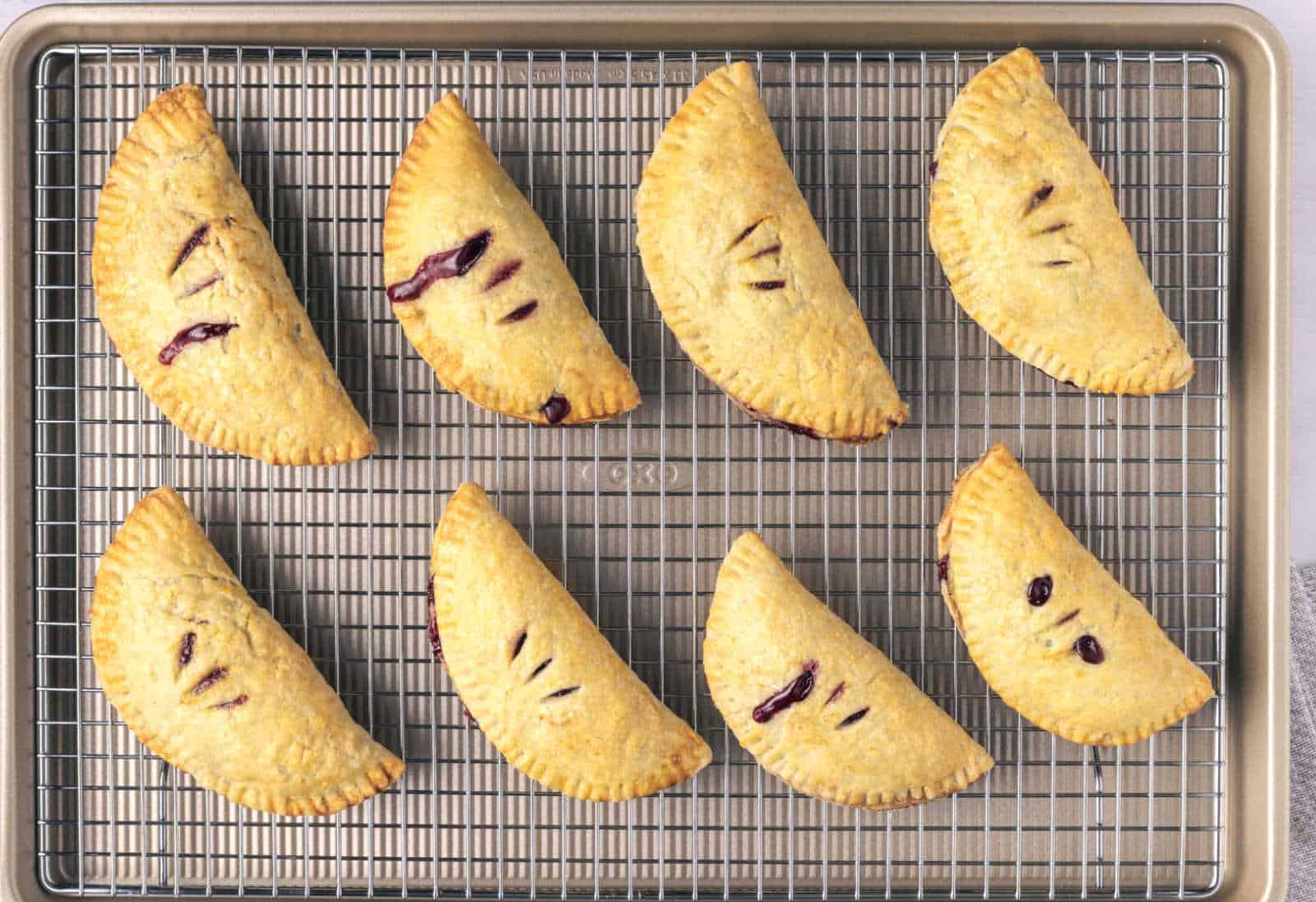 Eight golden-brown hand pies with vent slits are arranged on a wire rack over a baking sheet.
