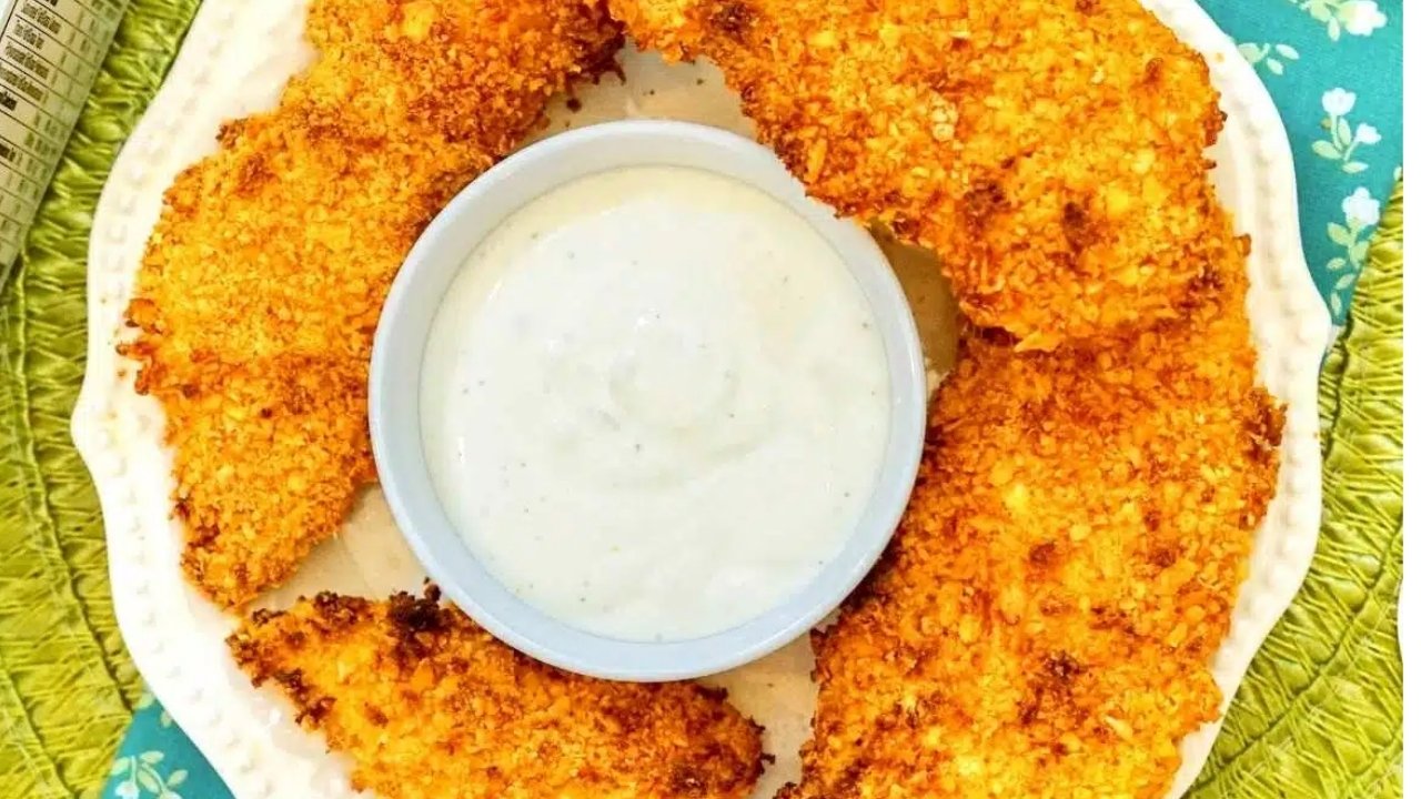 Five breaded chicken tenders arranged around a small bowl of white dipping sauce on a white plate.