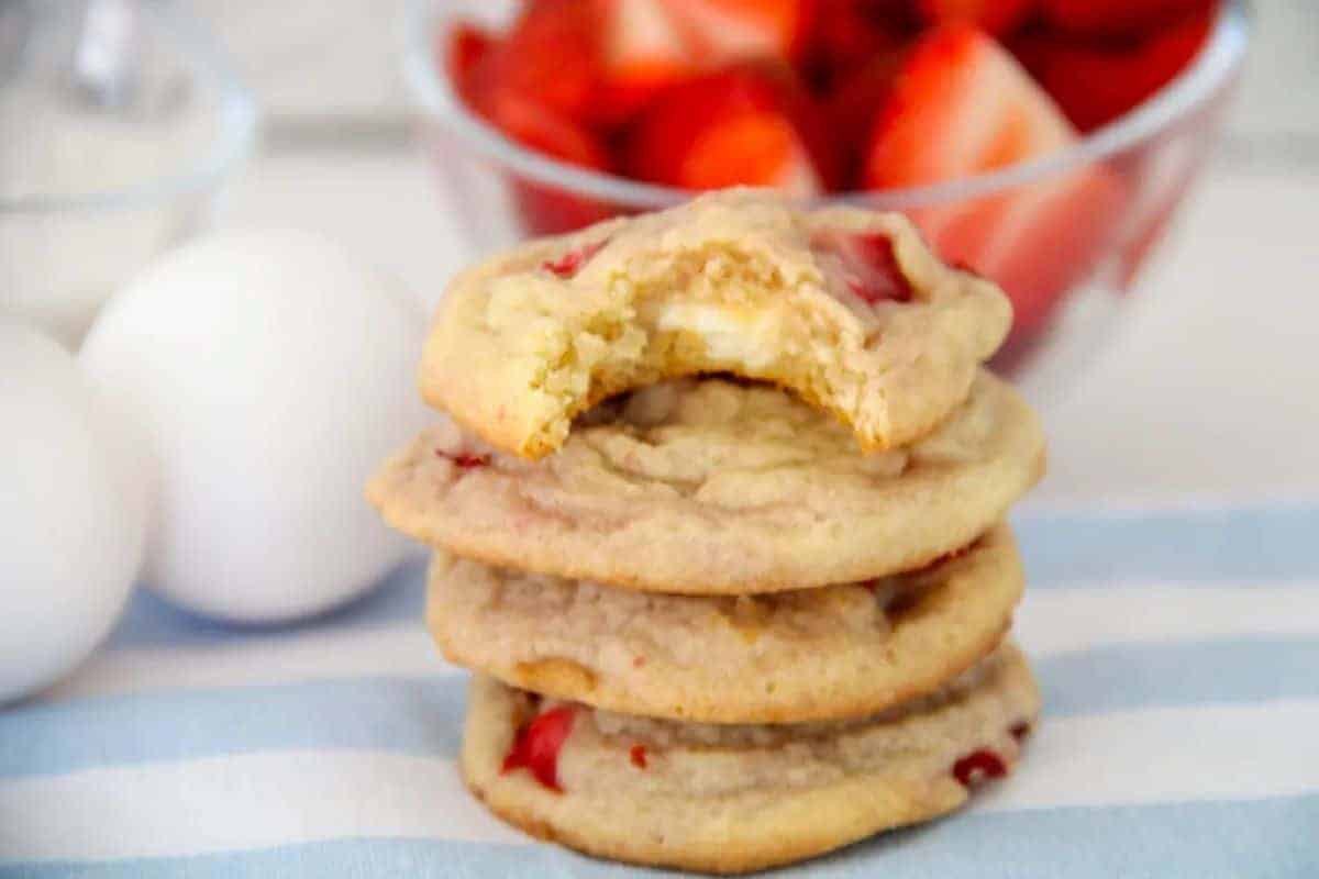 Three stacked cookies with visible strawberry pieces, one with a bite taken out, sit on a striped cloth next to eggs and a bowl of cut strawberries in the background.