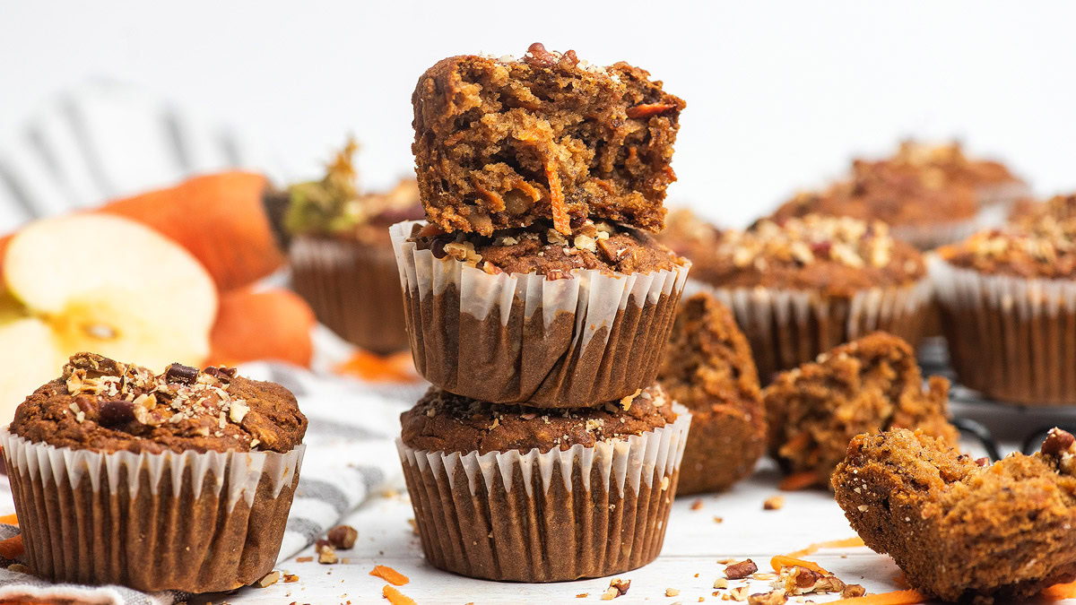 Three stacked muffins with one bitten on top, surrounded by more muffins, shredded carrots, and apple slices on a white surface.