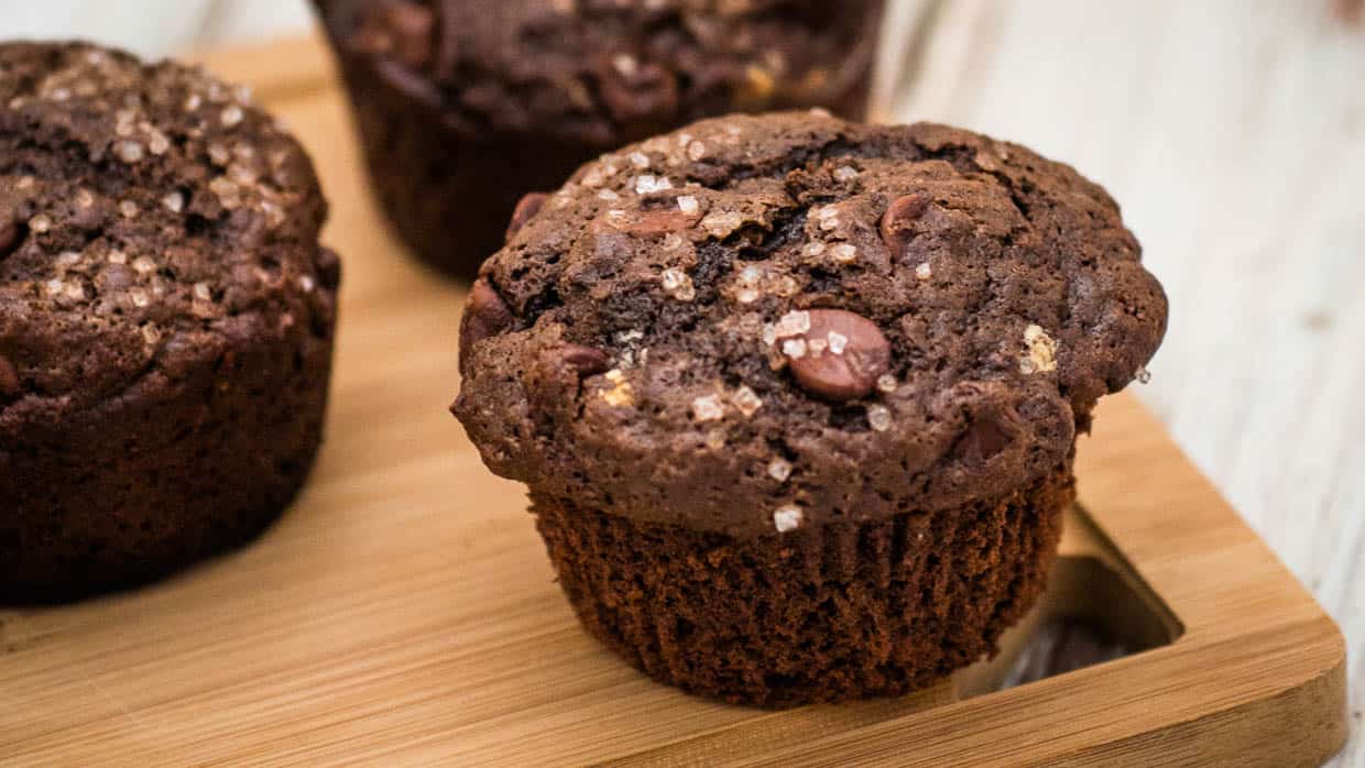 Close-up of a chocolate muffin with chocolate chips and sprinkled sugar on top, placed on a wooden board.