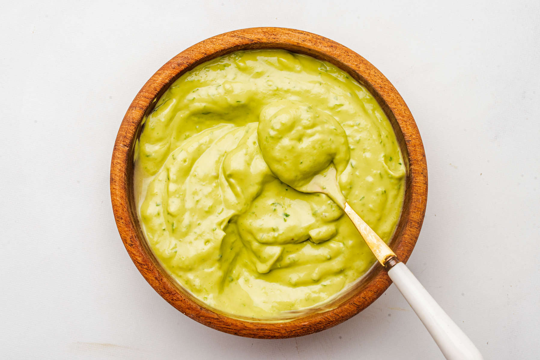 A wooden bowl filled with creamy green avocado sauce, with a spoon resting inside.