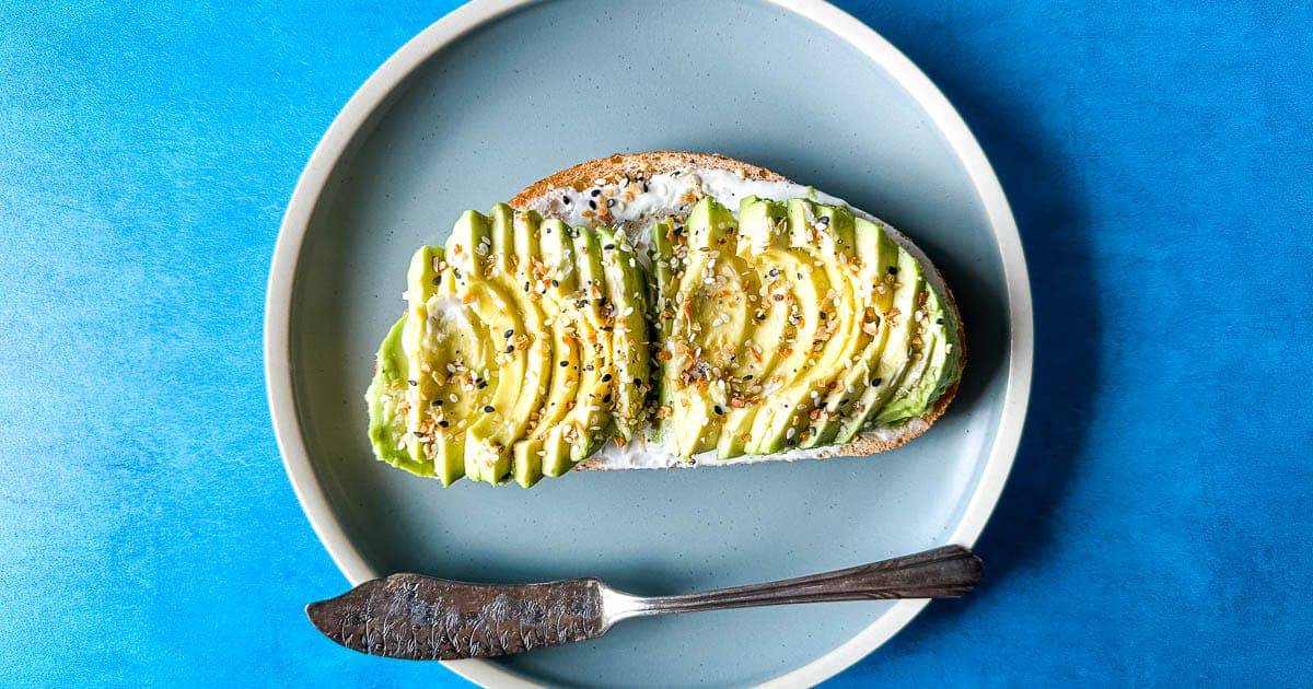 A slice of bread topped with cream cheese, sliced avocado, and seasoning sits on a round blue plate with a butter knife.