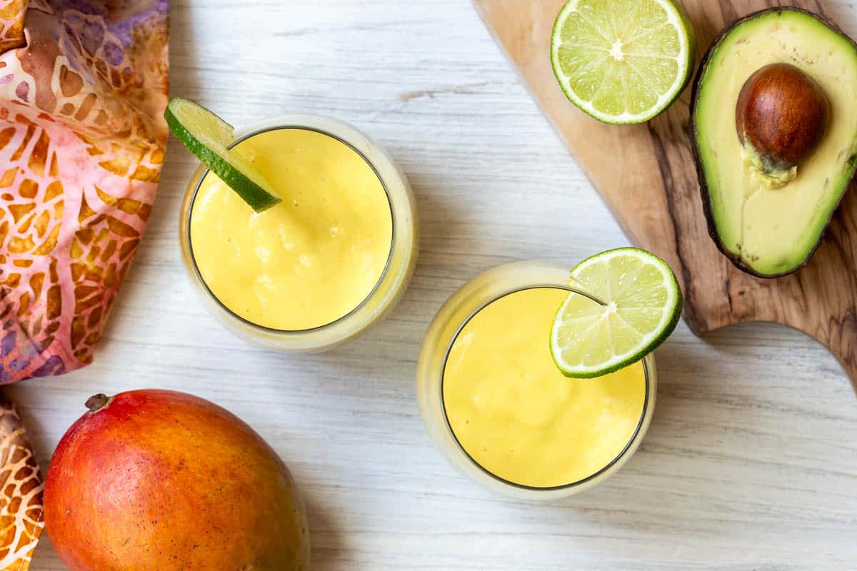 Two glasses of yellow smoothie with lime slices, next to a mango, sliced avocado, lime halves, and a colorful cloth on a white surface.