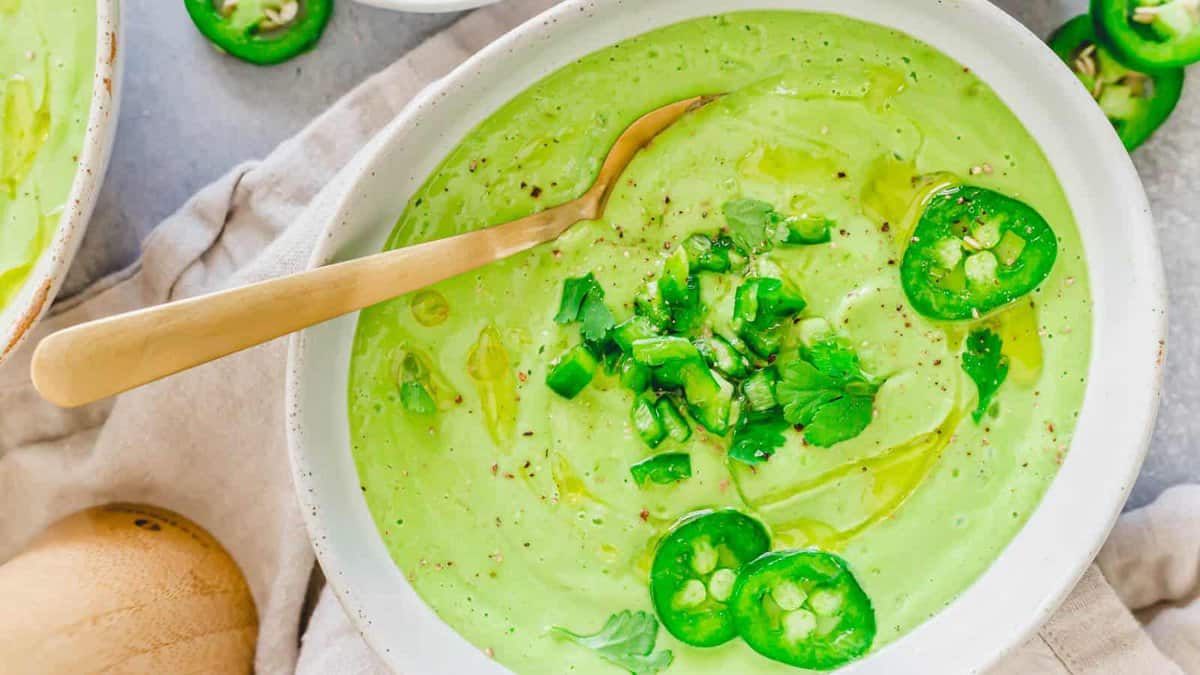 A bowl of creamy green soup garnished with sliced jalapeรฑos, chopped cilantro, black pepper, and a drizzle of oil, with a gold spoon resting inside.