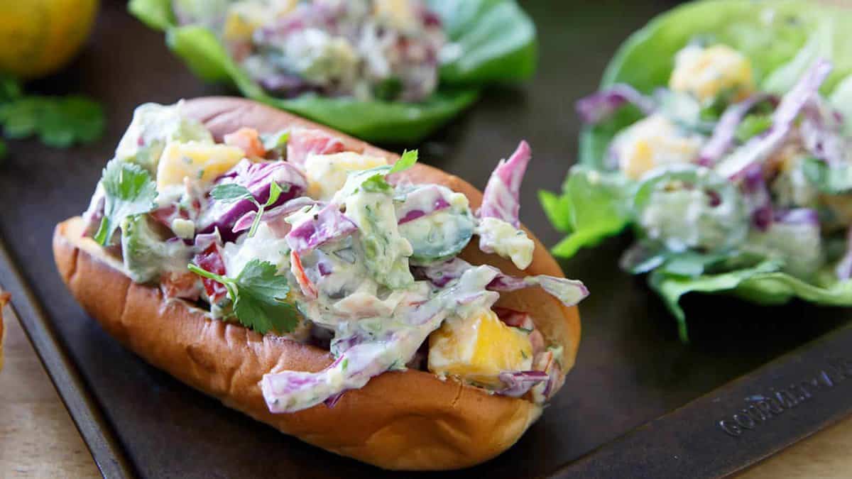 A sandwich bun filled with creamy mixed salad, including lettuce, purple cabbage, and mango pieces, with two lettuce-wrap versions in the background.