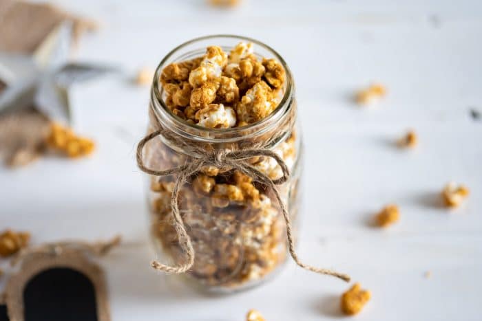 A glass jar filled with caramel popcorn, tied with a twine bow, sits on a white surface with scattered popcorn pieces around it.