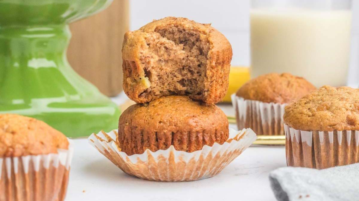 A stack of two banana muffins, with the top muffin partially eaten, sits on a white surface alongside more muffins and a glass of milk in the background.