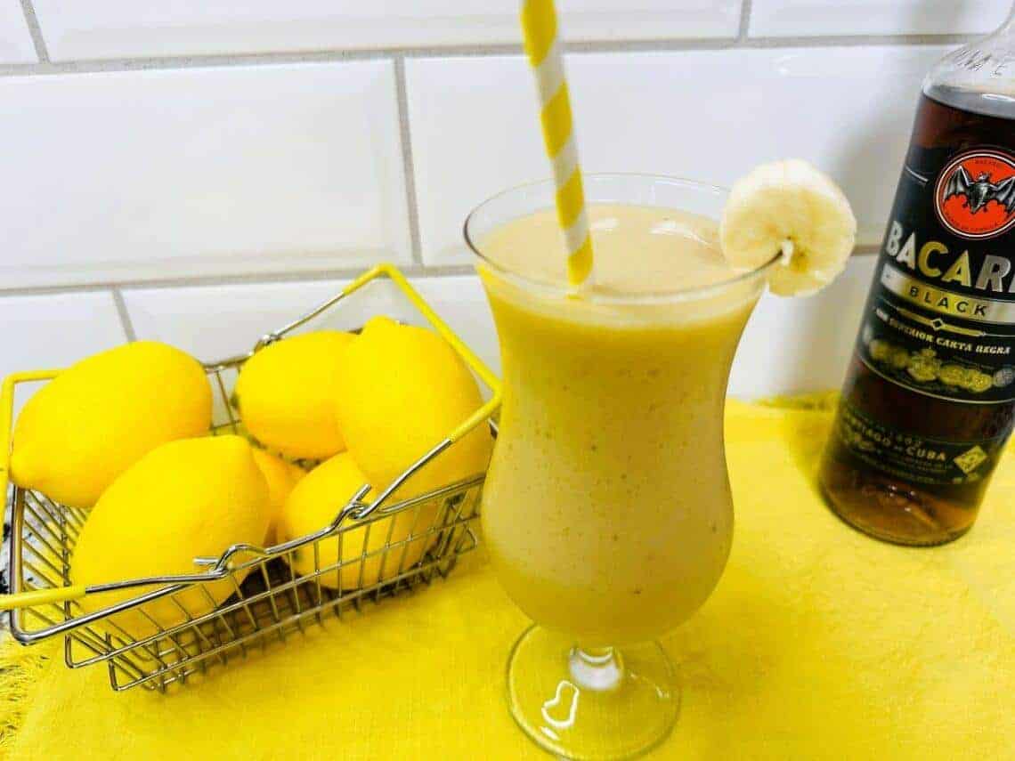 A banana smoothie garnished with a banana slice and striped straw sits on a yellow napkin beside a basket of lemons and a bottle of Bacardi rum.