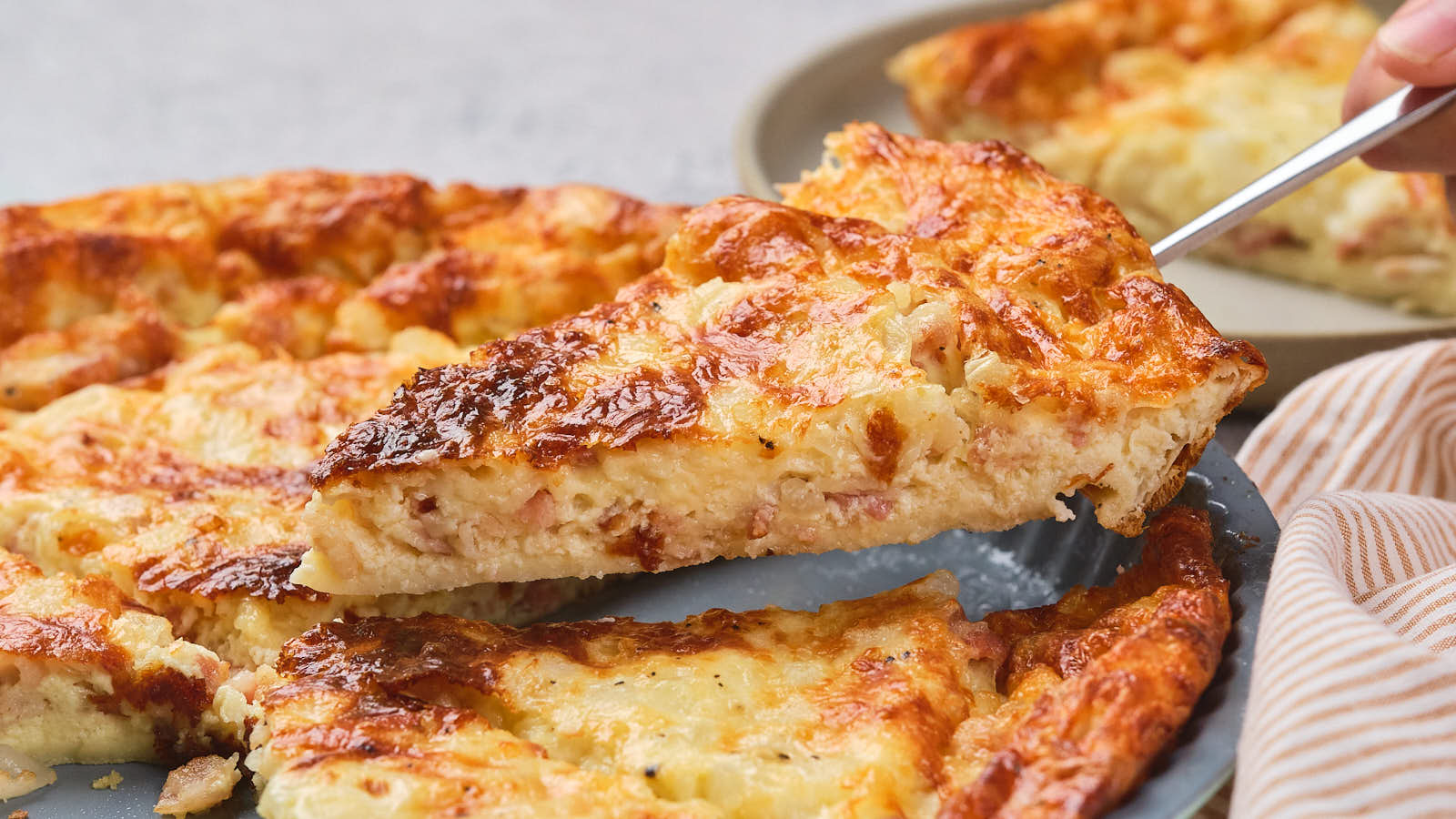 A close-up of a cheesy quiche with a slice being lifted from the dish, showing a golden-brown crust and creamy filling.