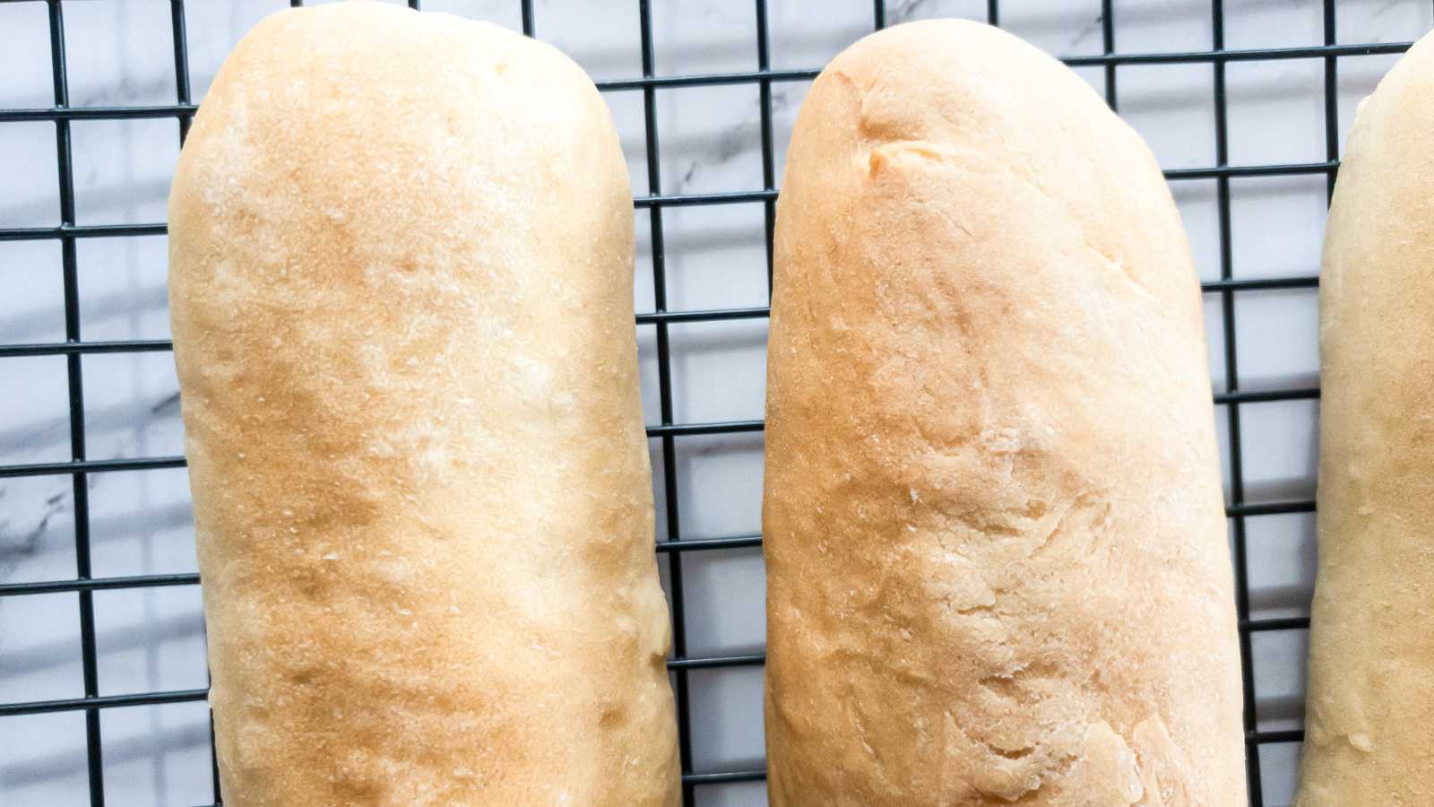 Two freshly baked hoagie rolls cooling on a black wire rack with a white marble background.