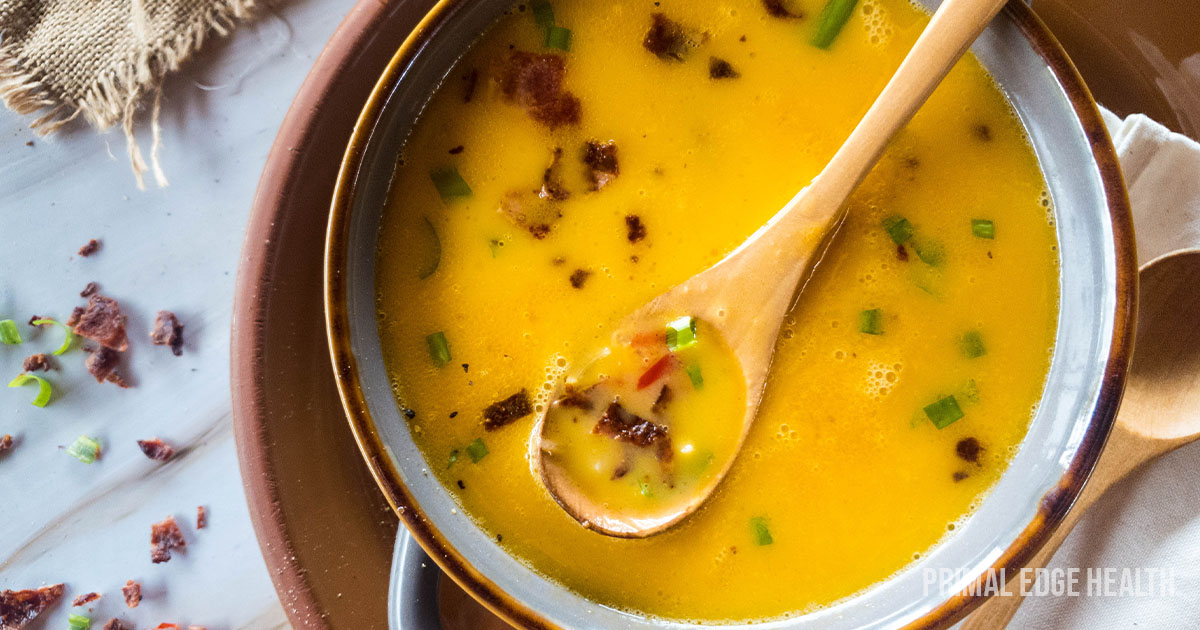 A bowl of yellow soup with chopped herbs and bacon bits, a wooden spoon inside, placed on a brown plate beside a wooden fork.
