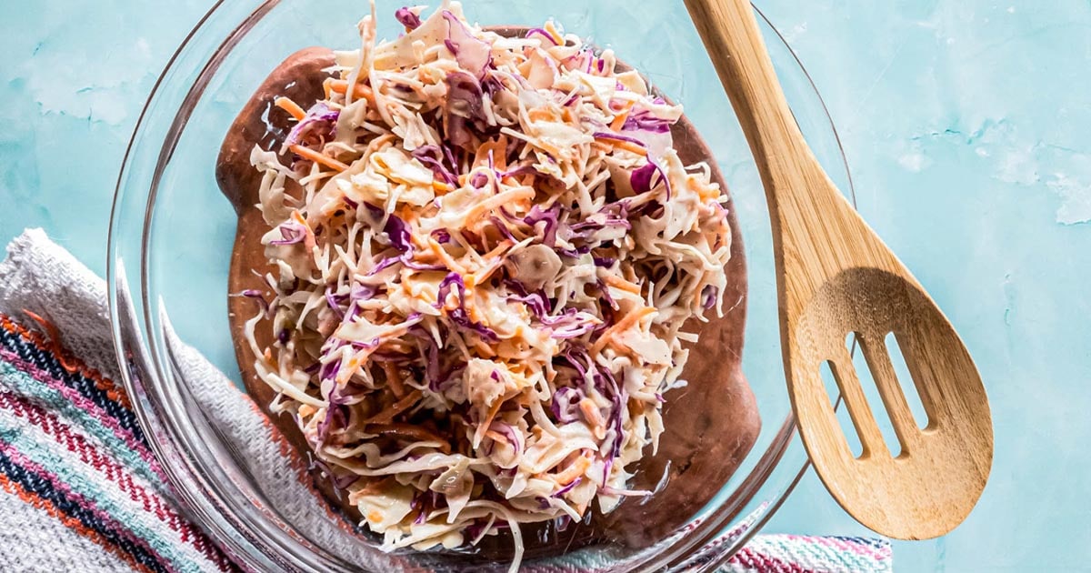 A glass bowl filled with creamy coleslaw made of shredded cabbage and carrots sits next to a wooden slotted spoon and a striped kitchen towel.