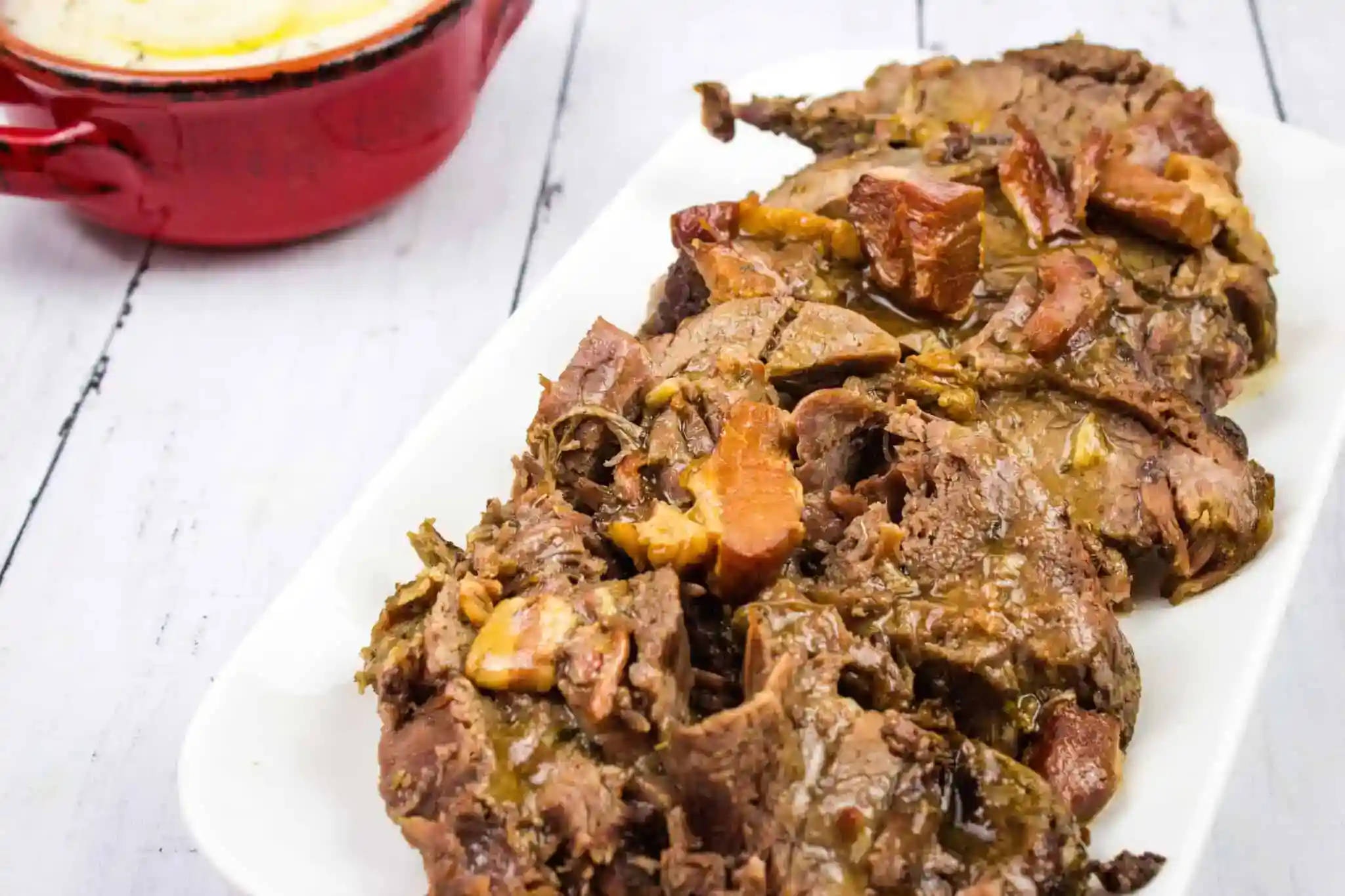Sliced pot roast with pieces of vegetables on a white plate, with a red casserole dish in the background on a light wooden surface.