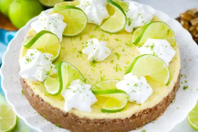 A key lime pie with graham cracker crust, topped with whipped cream, lime slices, and lime zest, displayed on a white cake stand.