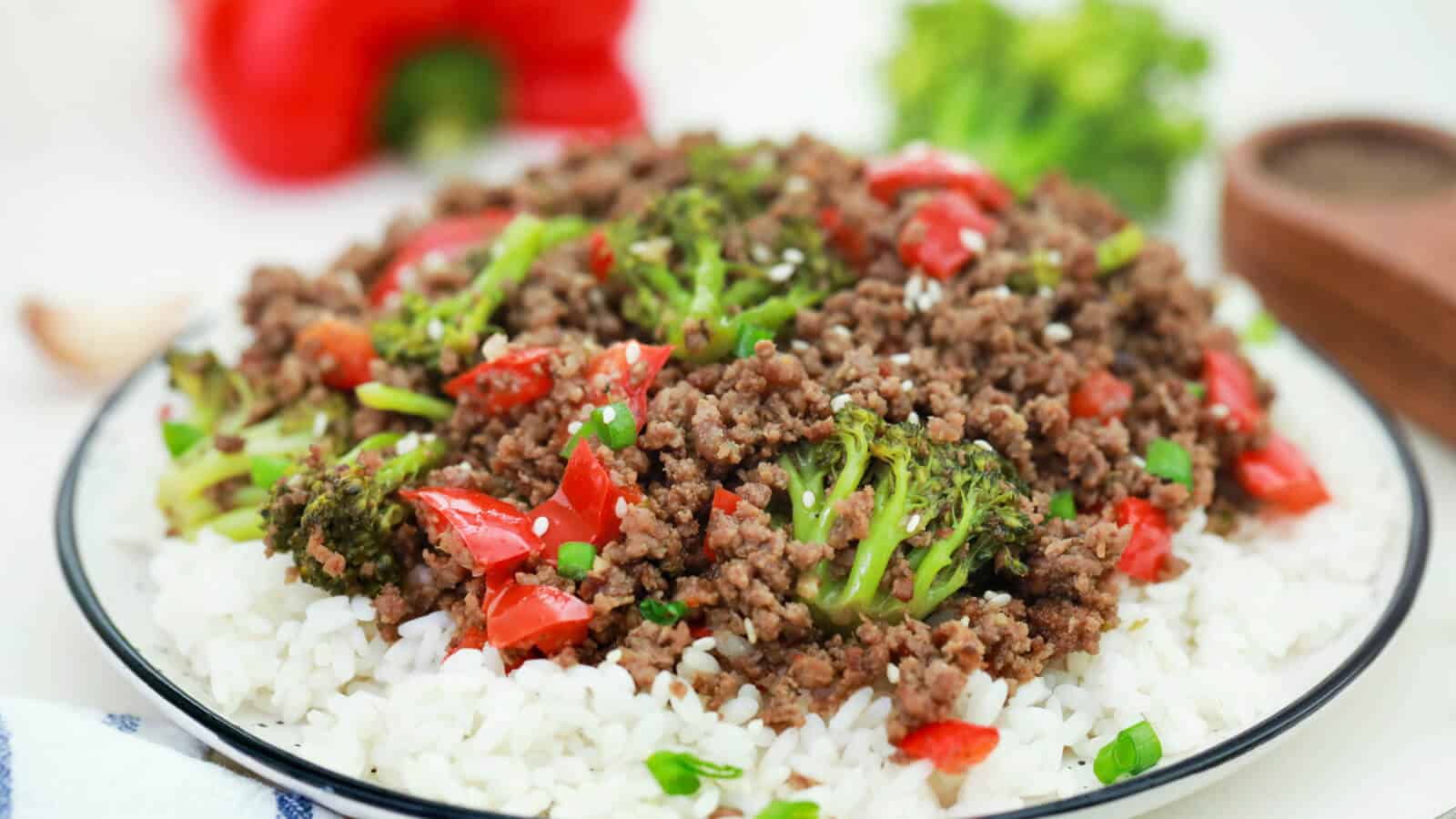A plate of white rice topped with cooked ground beef, broccoli florets, and diced red bell peppers.