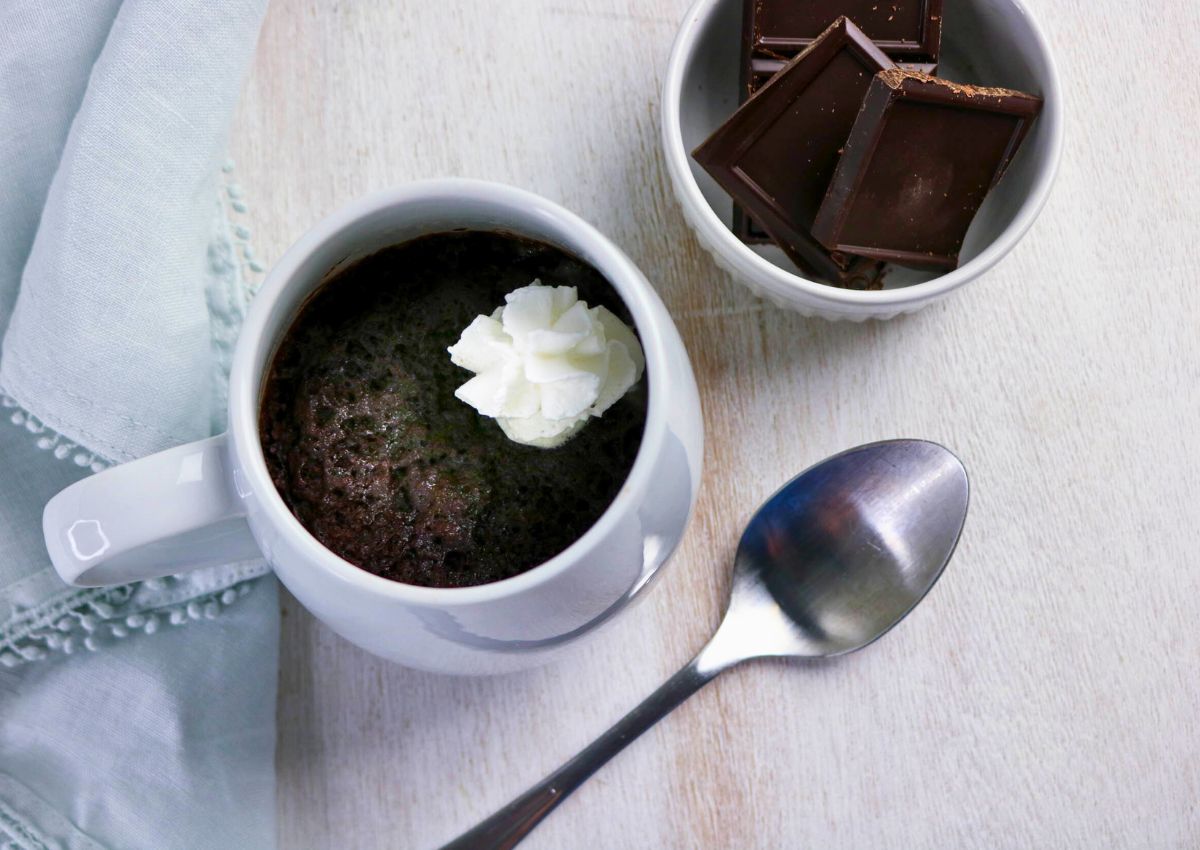 A chocolate mug cake topped with whipped cream sits beside a bowl of chocolate squares and a metal spoon on a light wooden surface.