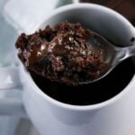 A spoon holds a serving of chocolate mug cake with melted chocolate, above a white mug containing the rest of the cake.