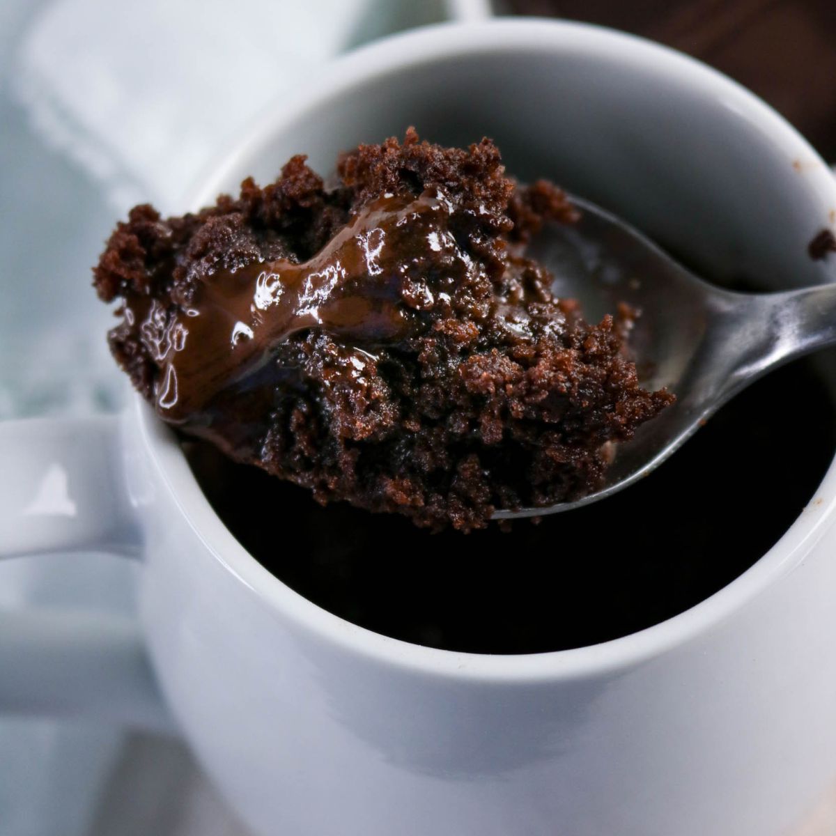 A spoon holds a serving of chocolate mug cake with melted chocolate, above a white mug containing the rest of the cake.