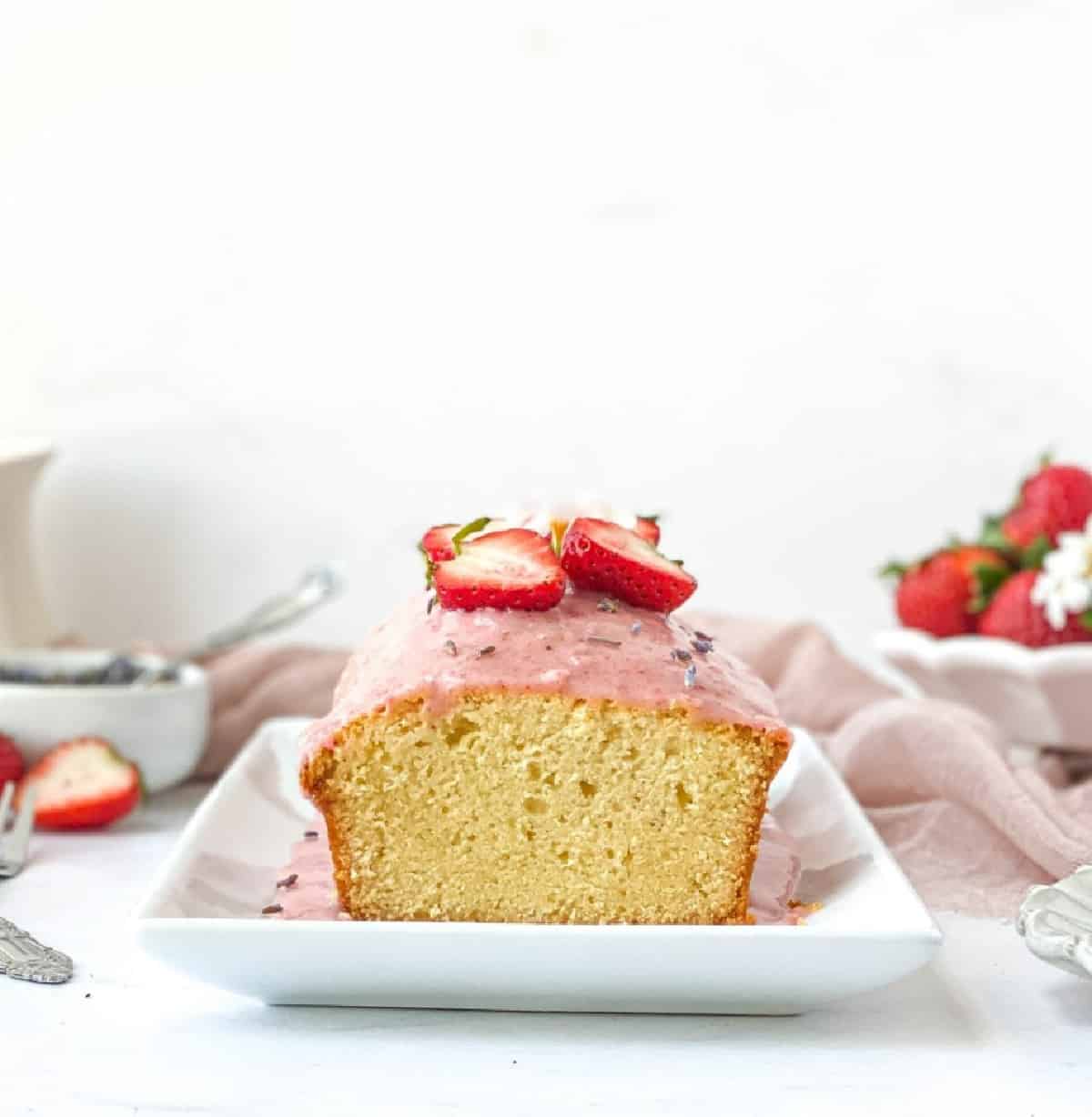 A loaf of pound cake with pink icing and sliced strawberries on top sits on a rectangular white plate, with strawberries and utensils in the background.